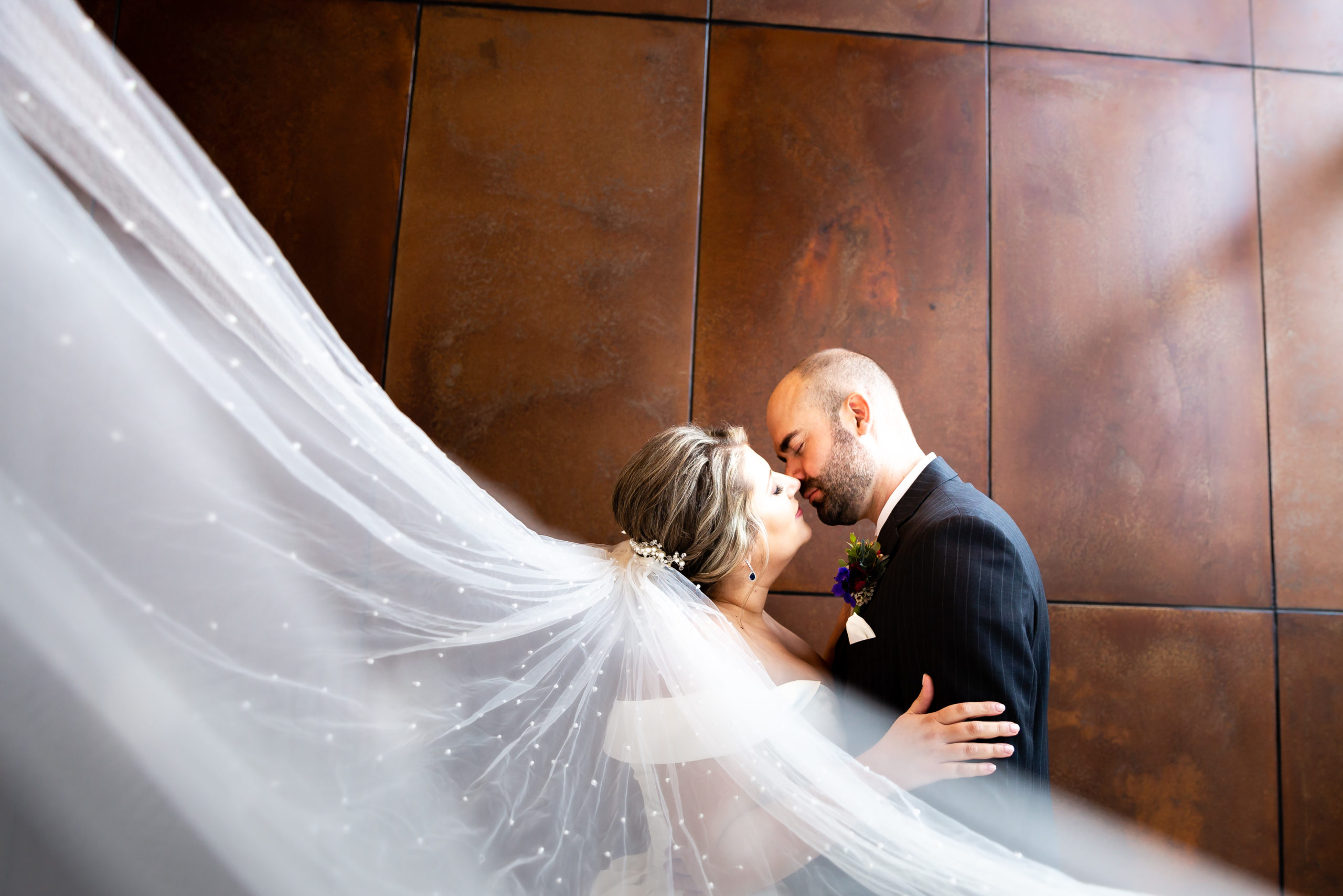 Portrait of bride and groom as her veil swoops towards the camera with a large copper paneled wall as the backdrop.