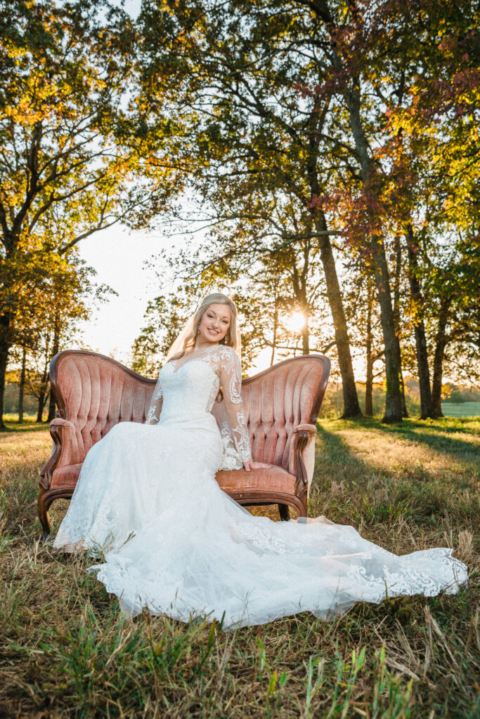 Bride seated on a vintage sofa outdoors during golden hour, wearing a long-sleeve lace wedding gown with fall trees glowing behind her.