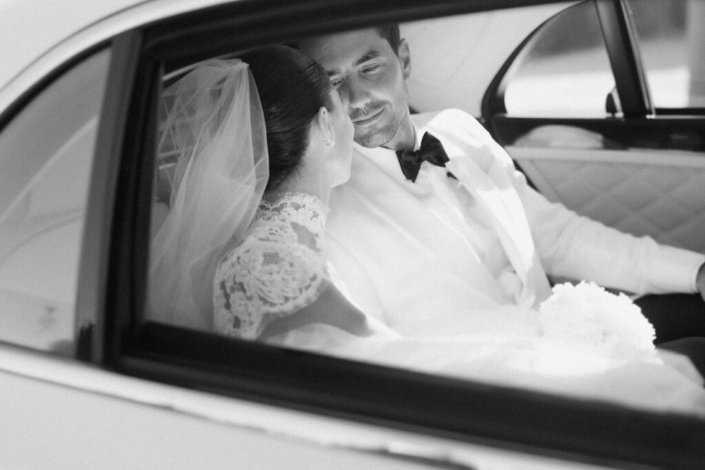 Black-and-white wedding photo of a bride and groom sitting closely in a classic car, captured in soft natural light with a timeless, cinematic feel.