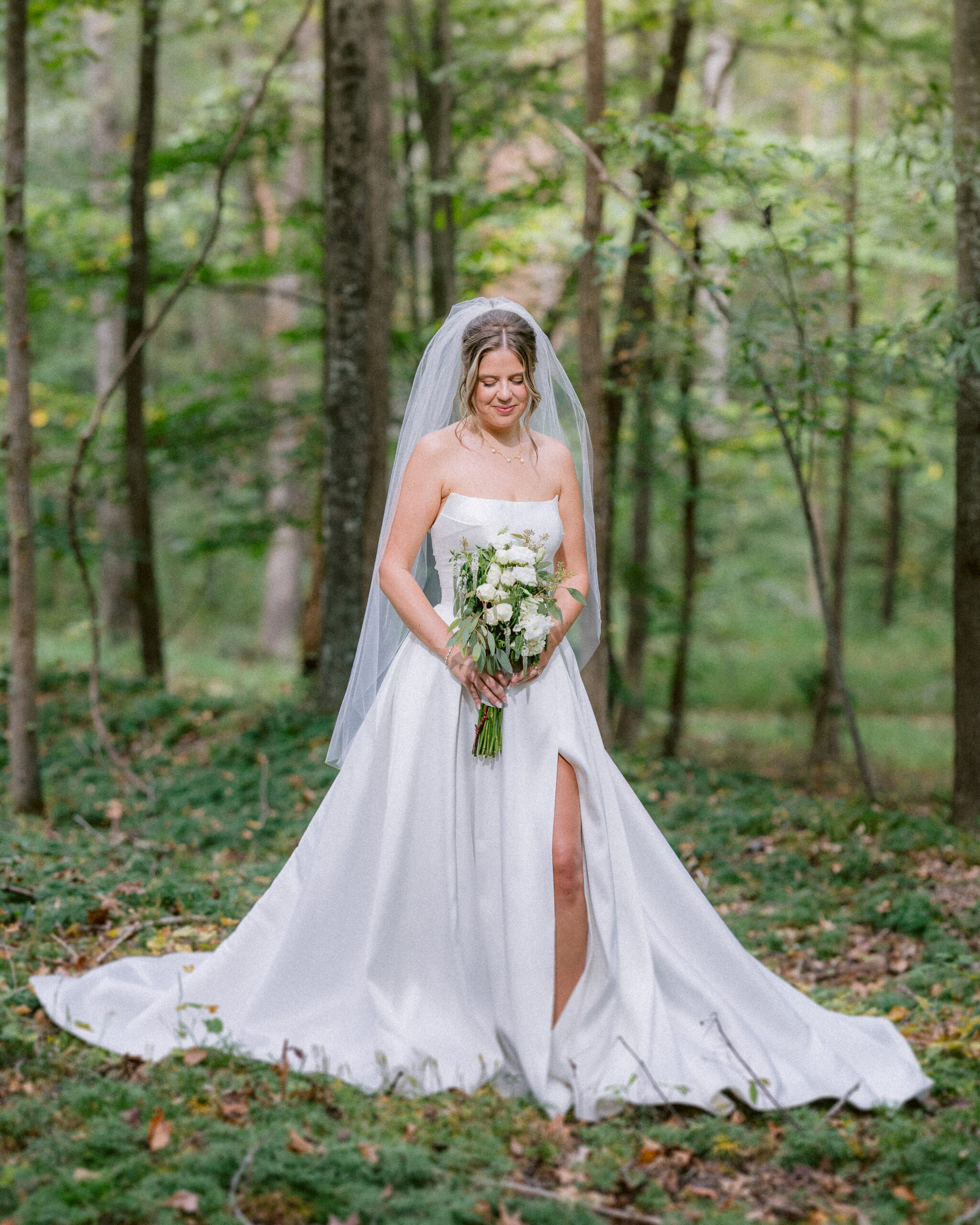 Bride standing alone in a wooded setting holding her bouquet, wearing a strapless wedding gown and veil, photographed in soft natural light.