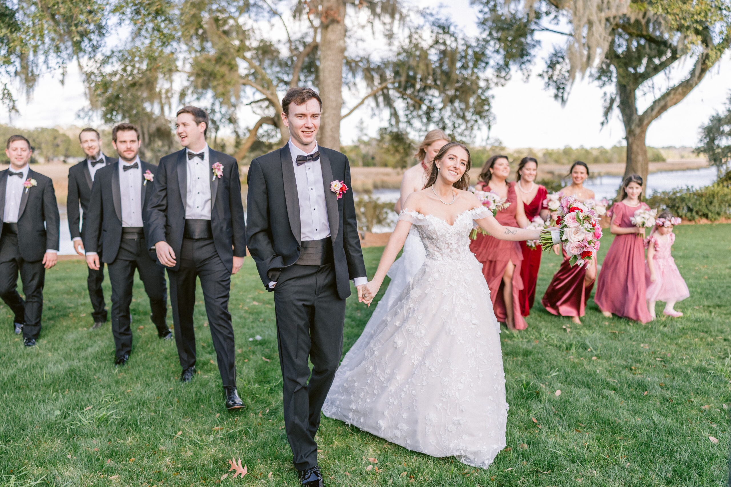 Bride and groom walking with their wedding party at an outdoor waterfront wedding venue, featuring oak trees, Spanish moss, bridesmaids in mauve dresses, and soft, natural daylight.