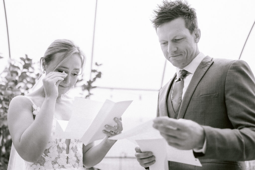 Bride wiping away tears as she reads her hand-written vows while the groom reads his during an intimate first-look before their wedding ceremony, photographed in black and white.