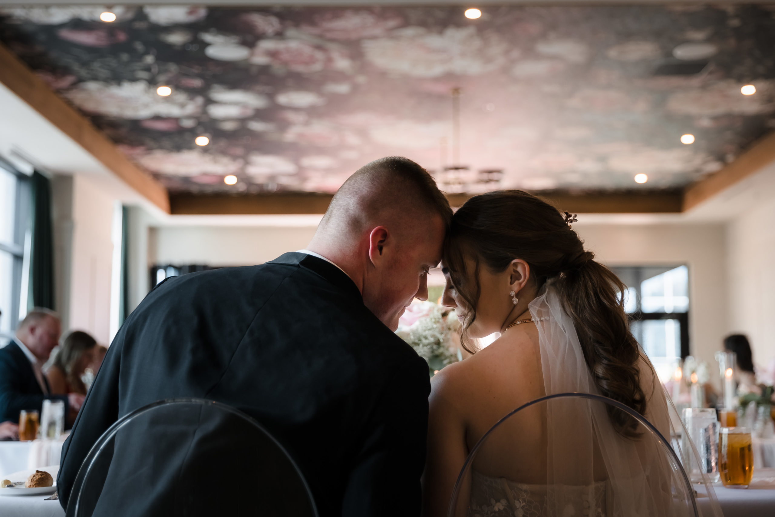 A bride and groom touch their foreheads together while prying for their first meal as husband and wife with a background of a floral mural painted on the ceiling.