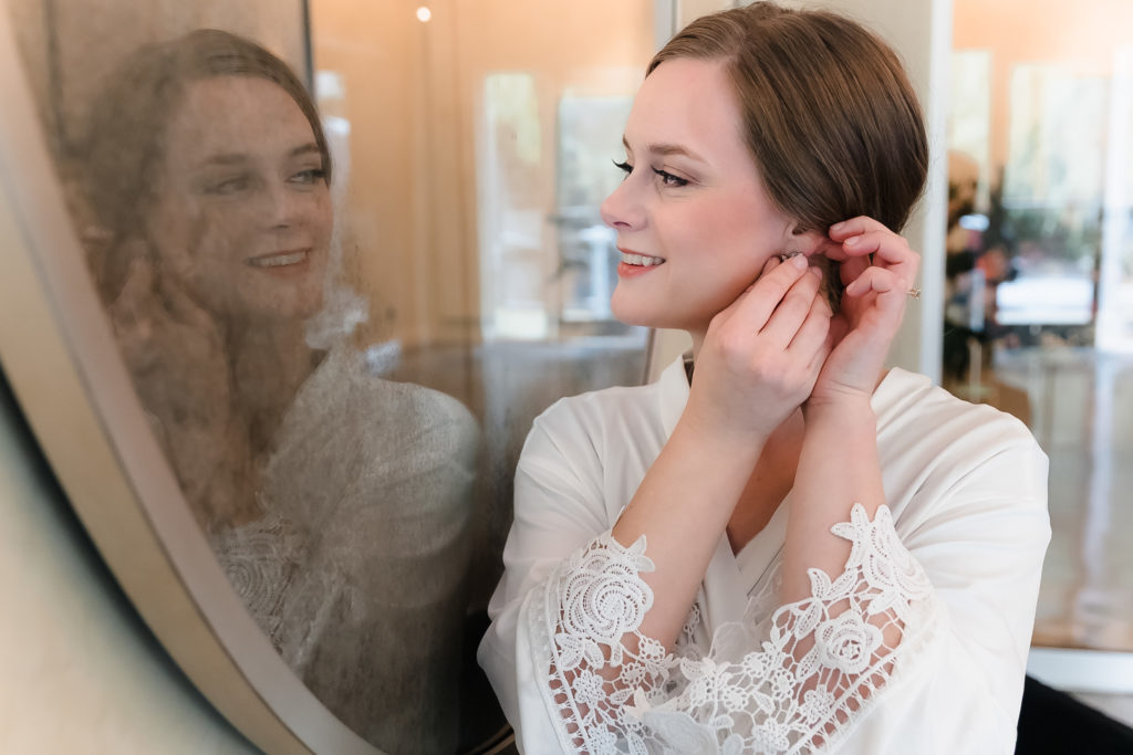 Bride looking into a decorative mirror to put on her earrings while wearing a silk lace robe.