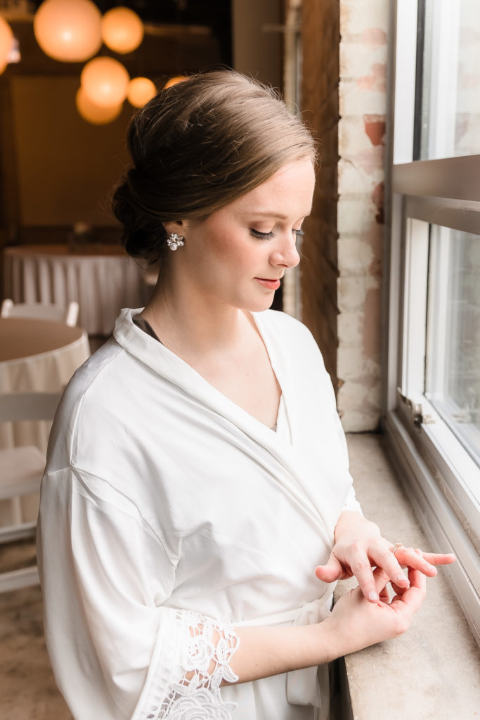 Bride standing in front of a window and looking at her wedding ring while in her robe getting ready.