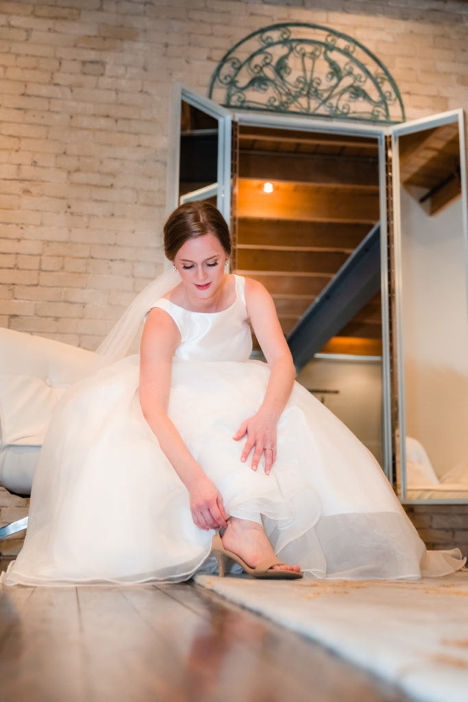 Bride leaning down to strap on shoes while wearing a large ball gown style wedding dress in the bridal suite of Zen - an elegant space for hire.