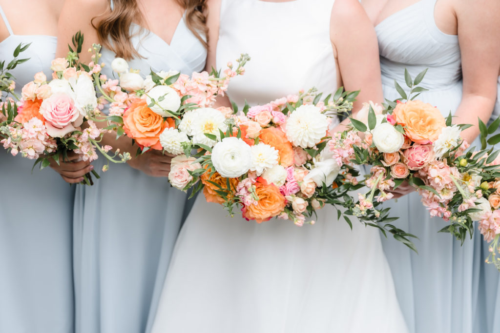 Closeup photograph of bride and bridesmaids bouquets that are filled with orange, white, and pink florals and lots of greenery.