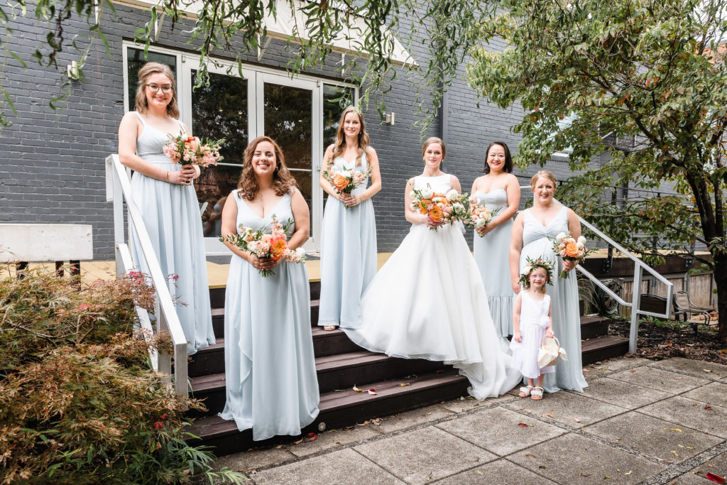 Bridesmaids and the bride standing on the steps with the flower girl. The bridesmaids are all wearing powder blue dresses and holding bouquets of white peonies, white dahlias, blush roses, and orange peonies.