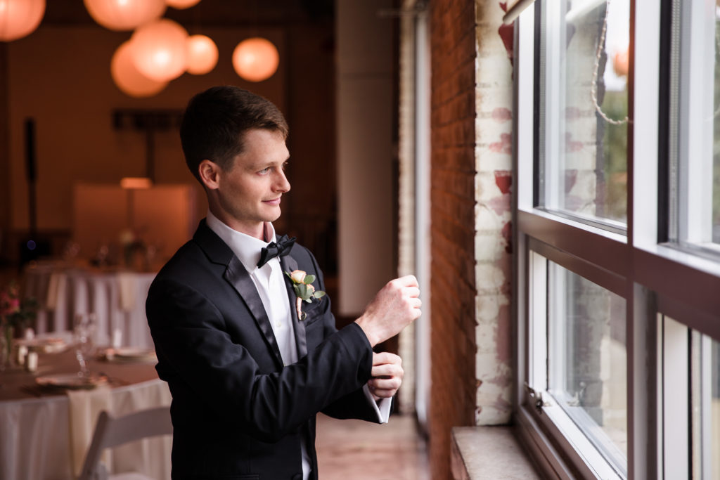 Groom adjusting the shirt cuffs of his tuxedo while looking out a large window.