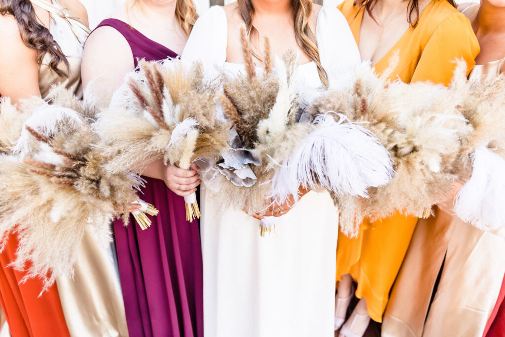 A boho bride and her bridesmaids wearing warm complimentary colors hold together their bouquets overflowing with pampas grass at a boho style wedding.