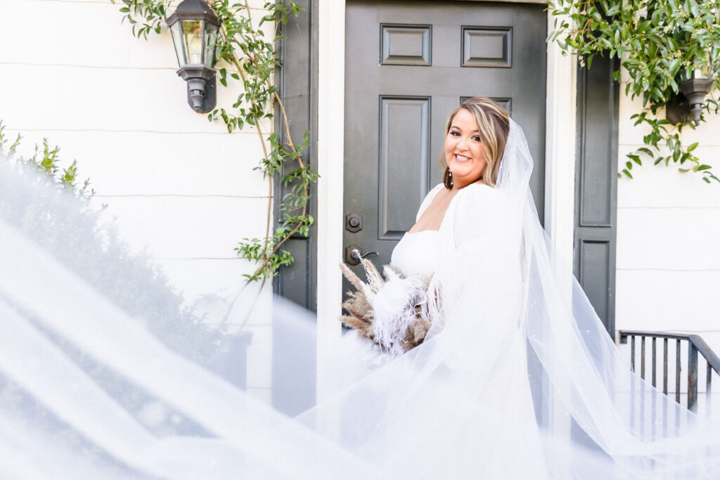 A brides veil swoops forward in the wind and leads into the bride as she holds her boho inspired bouquet with pampas grass in front of a a classic low carriage house facade with creeping ivy.