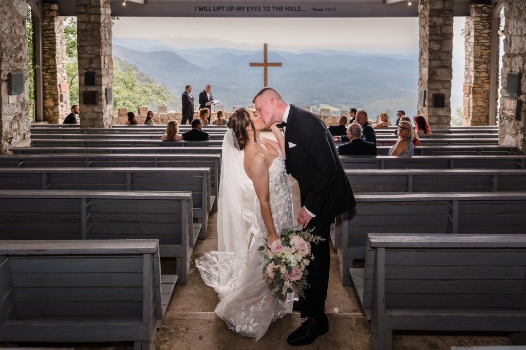 A groom dips and kisses his bride during their wedding recessional at Pretty Place Chapel with the cross and gorgeous mountain view in the background as their guests cheer for them.