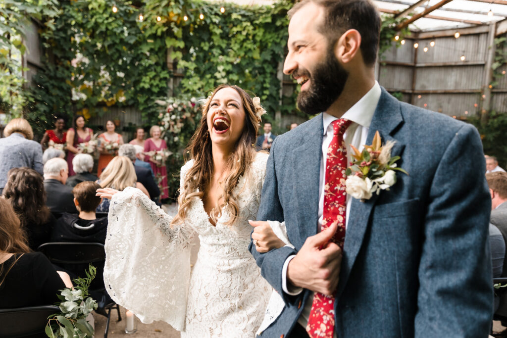 A bride and groom laugh and smile with joy as the walk arm in arm down the aisle after being married while wildflower seeds are thrown in the air by their guests. Her dress is fully lace with flowing sleeves and she wears white flower petals in her long hair. He wears a blue tweed suit with a bold red floral tie and a white rose boutonniere.