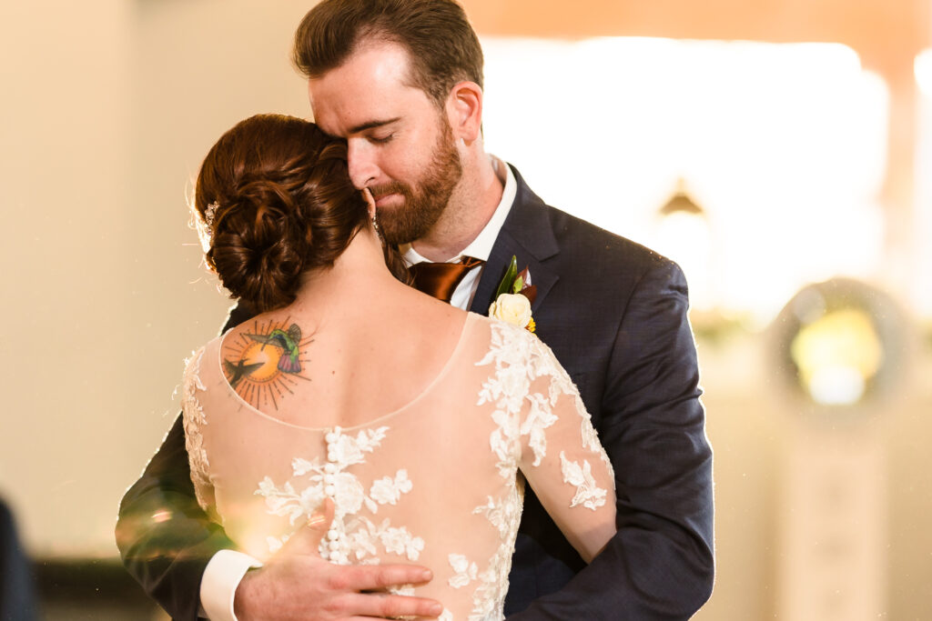 Newlyweds embrace during their first dance as golden light pours through a window behind them. 