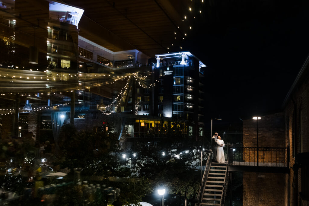 A bride and groom embrace during a moment of calm outside their wedding reception at Huguenot Mill & Loft with the Greenville riverwalk brightly lit behind them.
