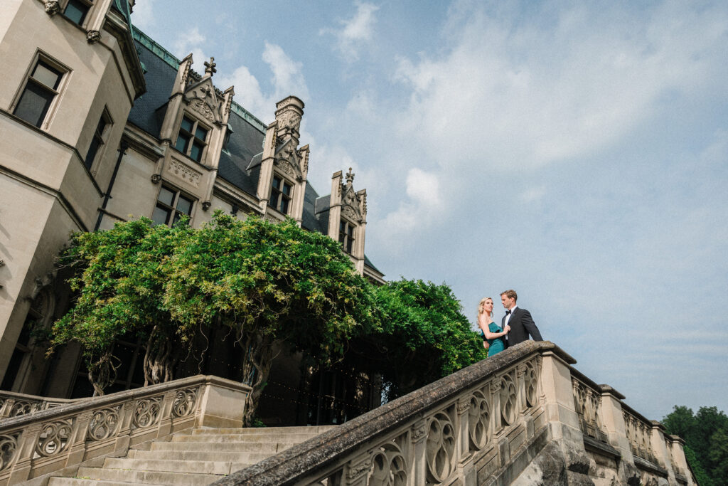 A couple embrace on the terrace stairs as the French renaissance style Biltmore Estate rises above them.