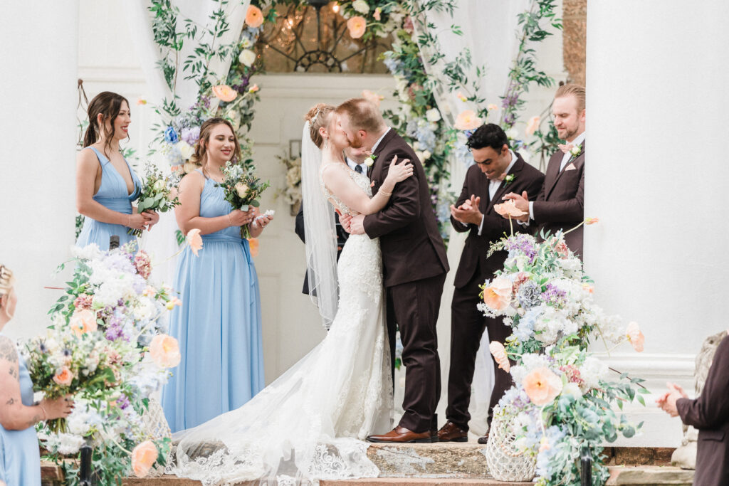 Bride and grooms first kiss surrounded by dripping florals and their bridal party.