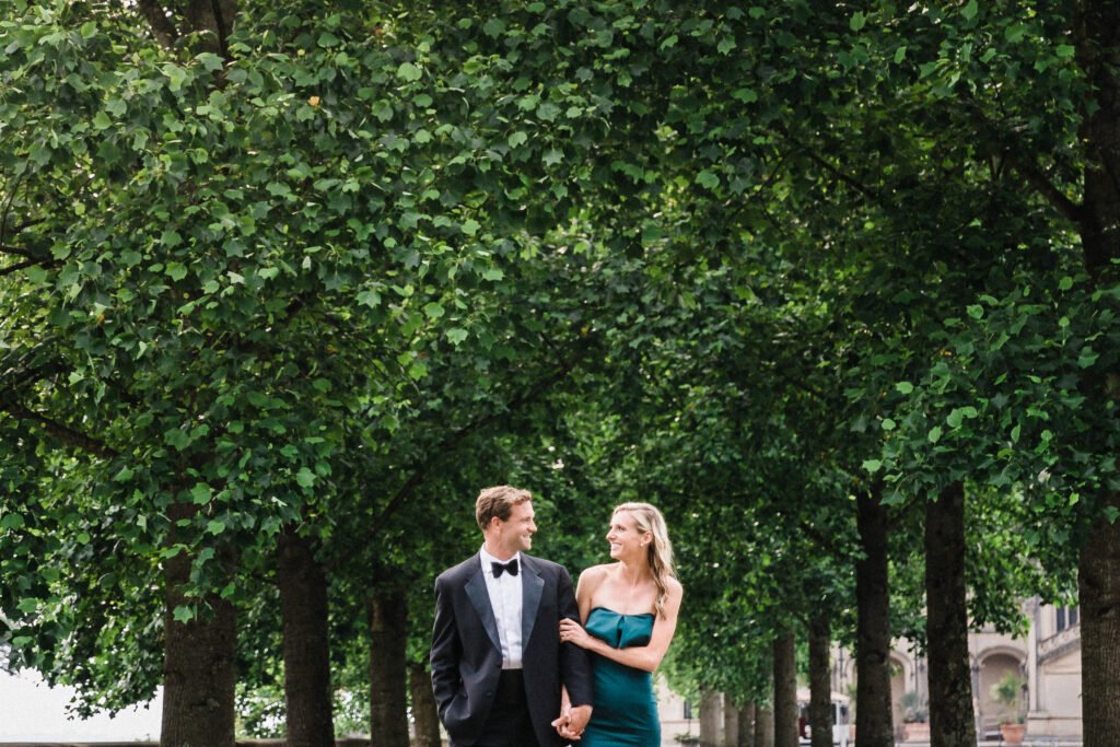 An engaged couple wearing an emerald green off the shoulder gown and classic black and white tuxedo walk through an allée of trees at the Biltmore estate.