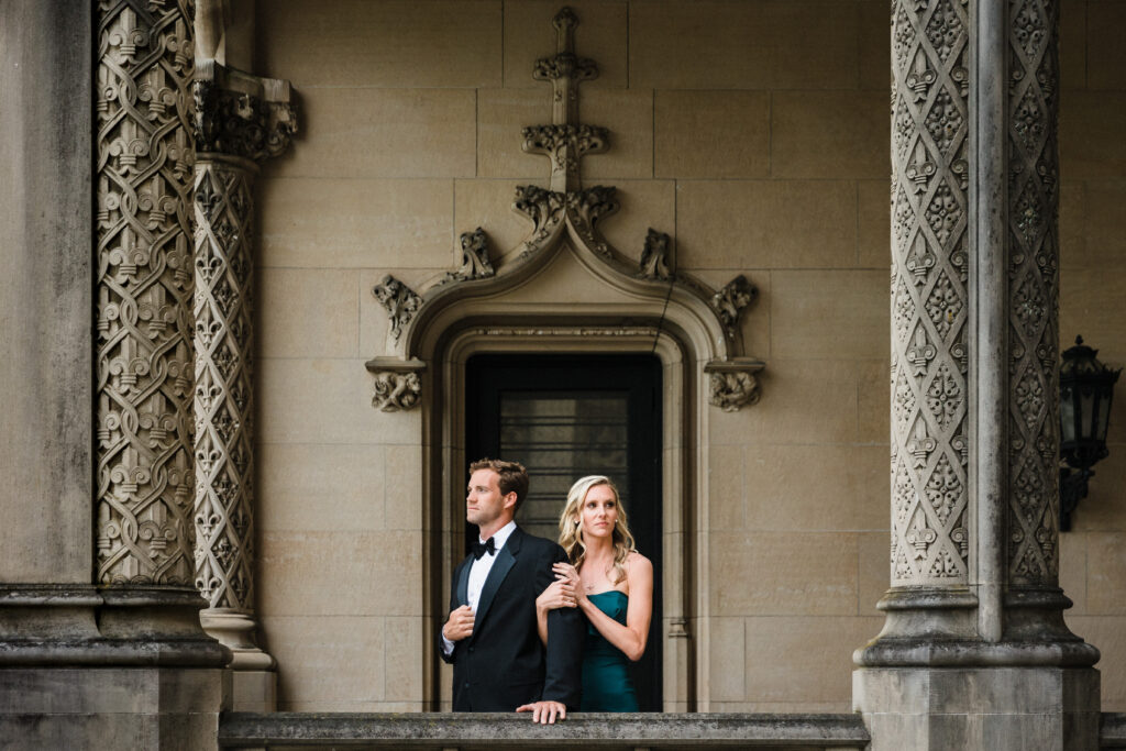 A sophisticated engaged couple wear an emerald green cocktail gown and classic black tuxedo and bowtie while framed by the châteauesque architecture of the Biltmore Estate.