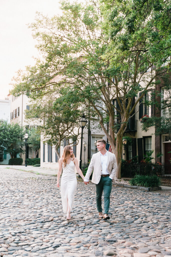 An engaged couple walk the cobblestone streets of Charleston wearing neutral tones that match the natural beige and greens of the historic city.