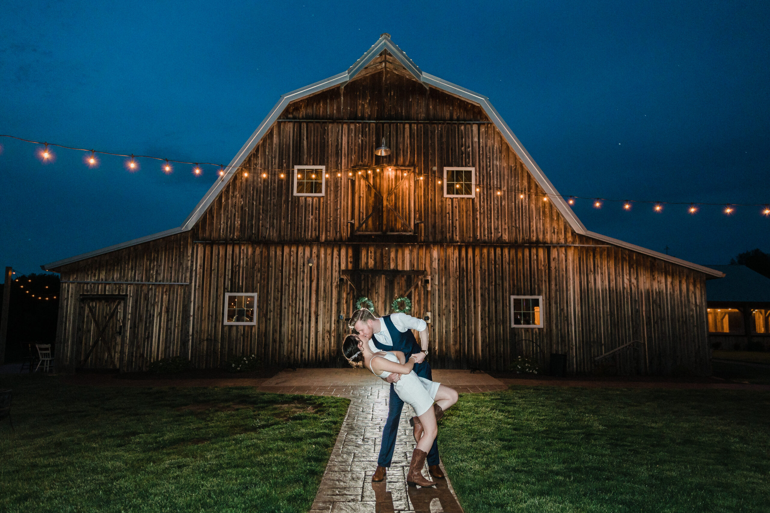 A couple dips and kisses in front of a rustic barn wedding venue with string lights overhead.