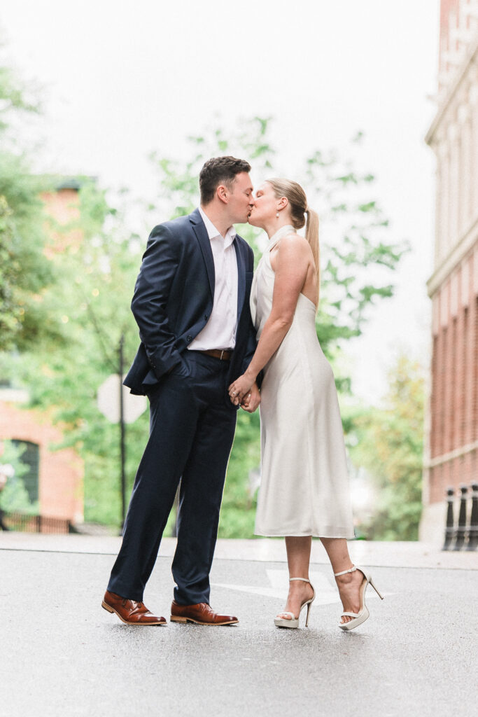 An engaged couple lean in for a kiss while walking in the city. She is wearing a neutral cream colored dress with pearl earrings and white heels while he wears a navy blue suit with brown belt and oxfords.