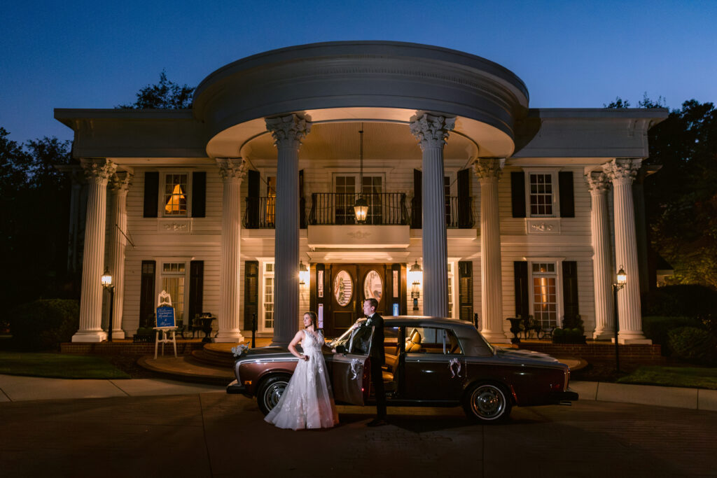 A portrait of a couple with a Rolls Royce in front of the stunning columns and front facade of the Grand Holland Estate during blue hour the couple stands in front of the car in a floral designed bridal gown and full tuxedo attire.