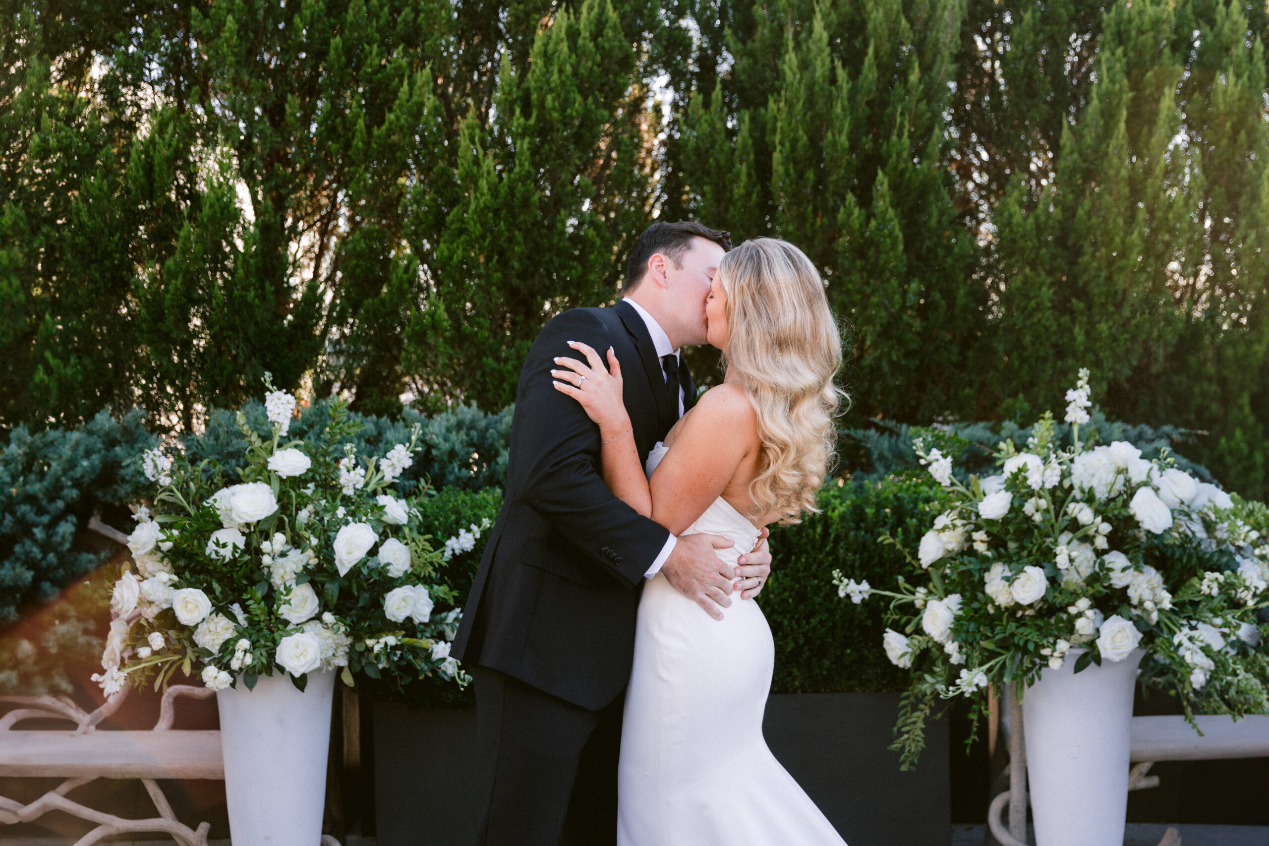 A bride and groom share their first kiss as husband and wife during their wedding ceremony at Avenue in downtown Greenville, SC in front of a wall of florals and greenery.