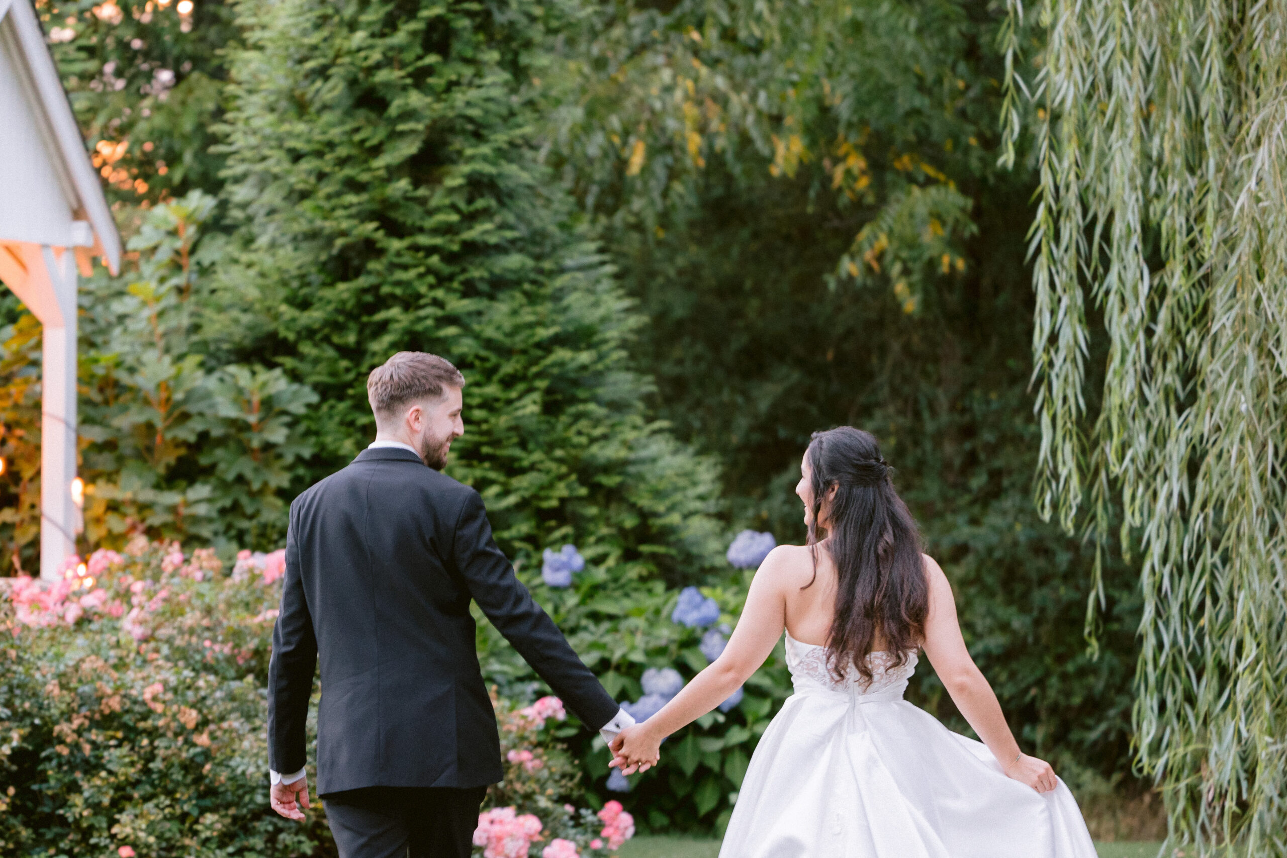 A bride and groom hold hands and walk into the garden of Aurora Farms towards a beautiful hydrangea and weeping willow tree.