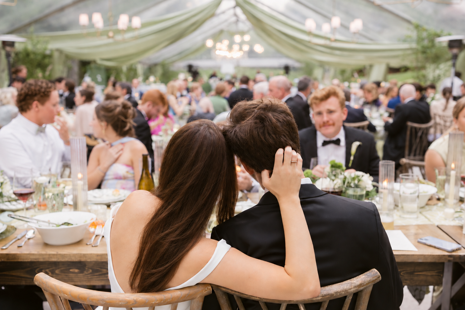 Bride resting her head on the groom's shoulder during dinner, with guests seated throughout a clear-top tent behind them