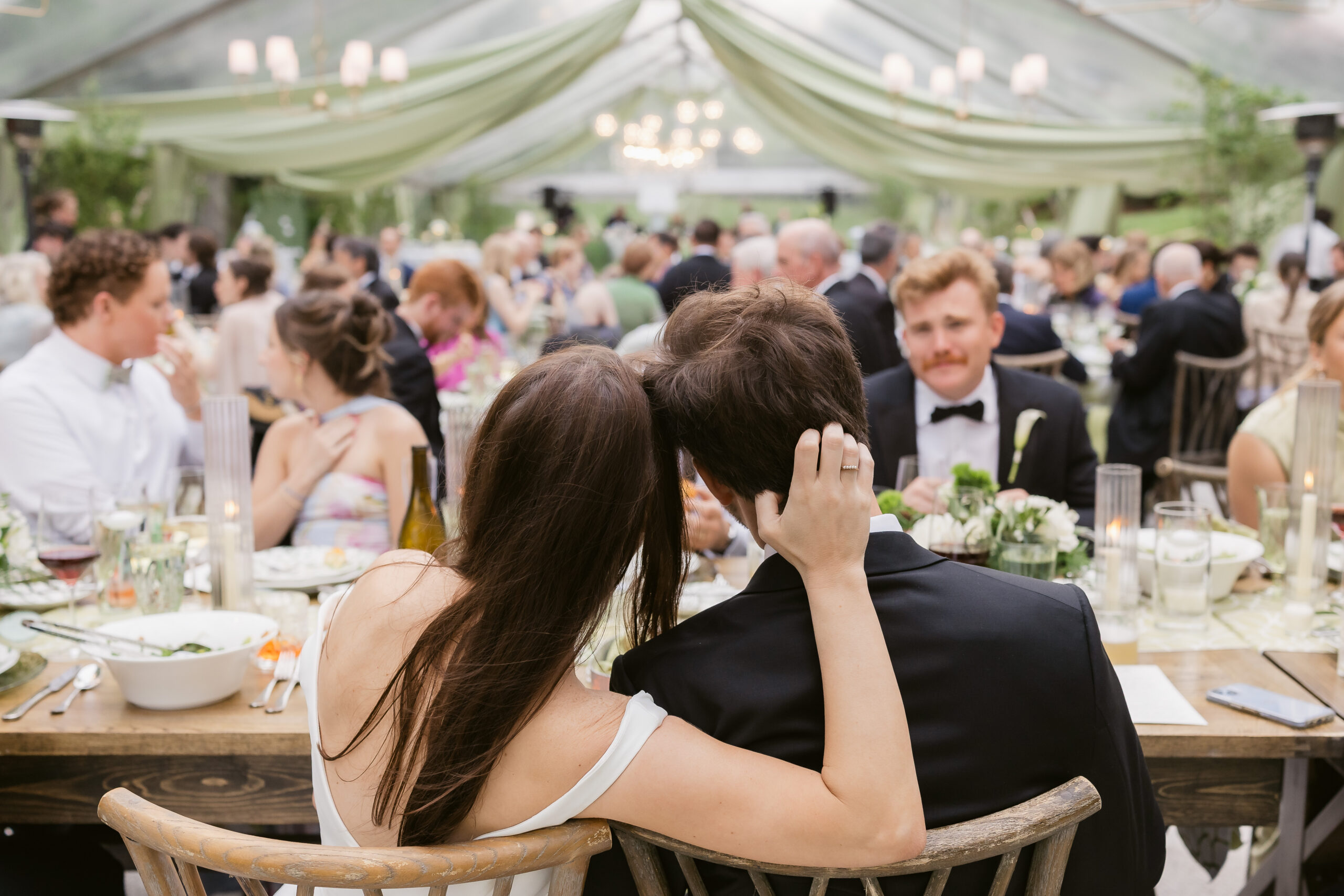 Bride resting her head on the groom's shoulder during dinner, with guests seated throughout a clear-top tent behind them