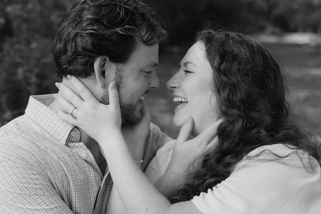 Black-and-white engagement portrait of a couple laughing together and showing an engagement ring